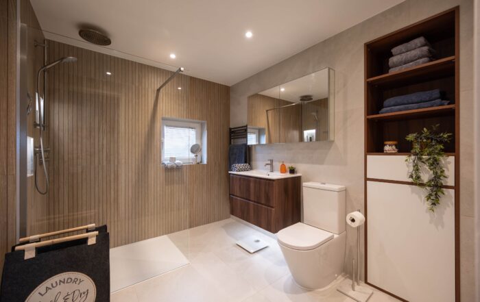 Full room view of Bermondsey main bathroom showing walk-in shower with glass panel and ceiling rainfall head, wall-hung walnut vanity, open shelving unit and large format neutral floor tiles
