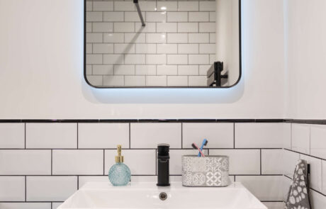 bermondsey-second-bathroom-illuminated-mirror-dark-vanity-metro-tiles.jpg Close-up of second bathroom vanity with backlit rounded mirror, black matte tap and dark grey cabinet unit against white metro tiles