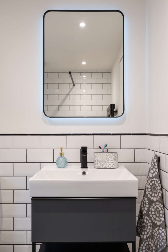bermondsey-second-bathroom-illuminated-mirror-dark-vanity-metro-tiles.jpg Close-up of second bathroom vanity with backlit rounded mirror, black matte tap and dark grey cabinet unit against white metro tiles