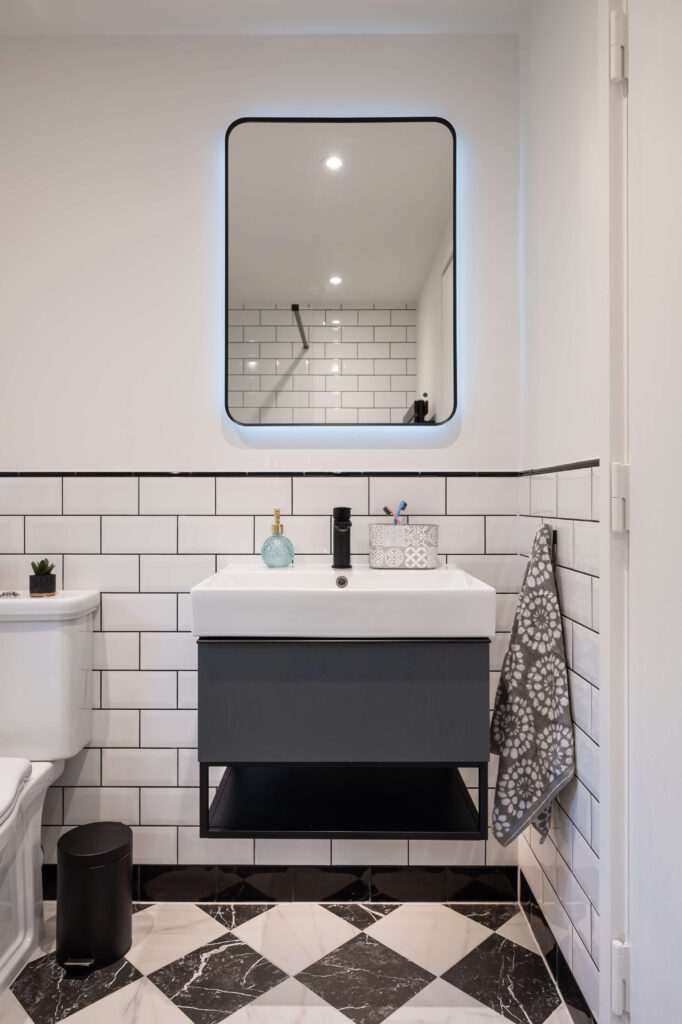 bermondsey-second-bathroom-metro-tiles-dark-vanity-illuminated-mirror.jpg Close-up of second bathroom vanity with backlit rounded mirror, black matte tap and dark grey cabinet unit against white metro tiles