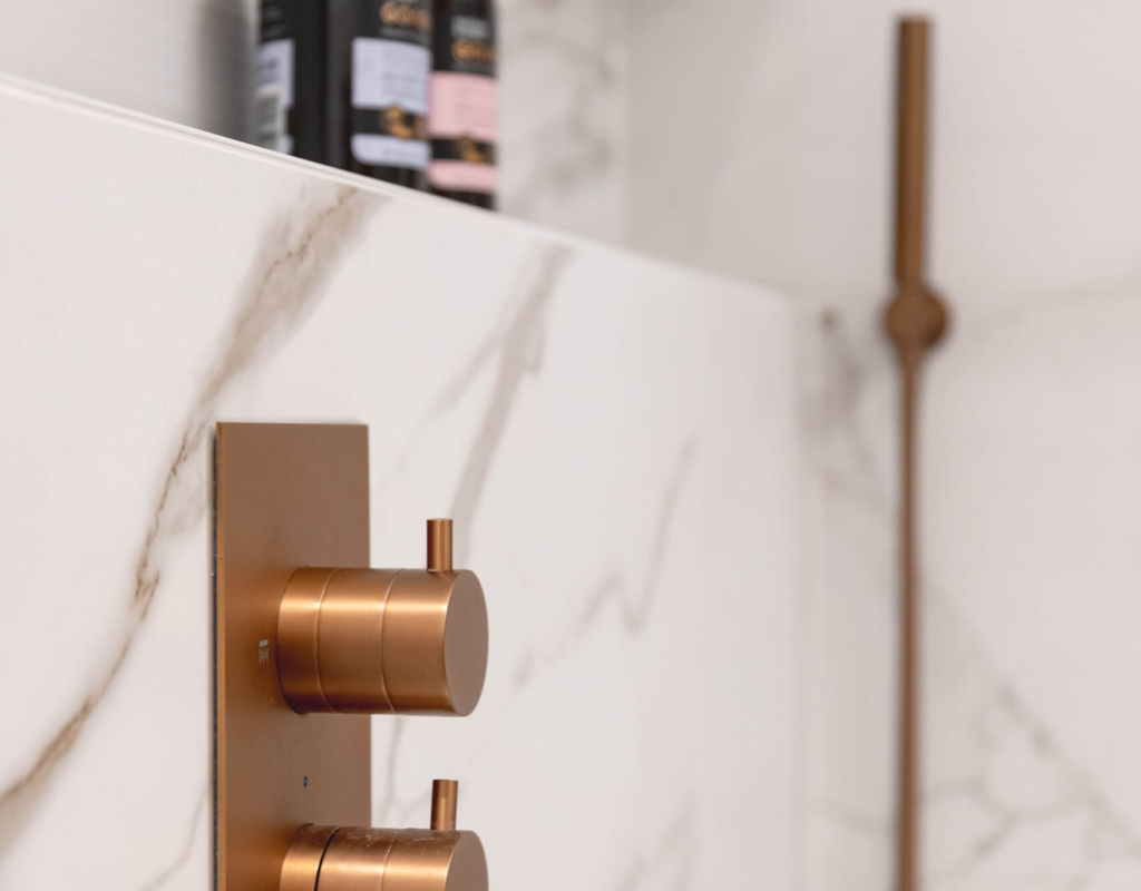 Close-up of brushed copper thermostatic shower controls set into marble-effect tiled wall with recessed niche and handheld shower rail beyond