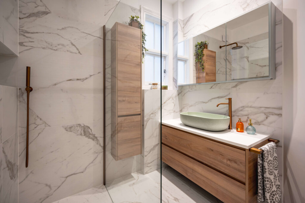 hugo-oliver-bathroom-full-view-shower-vanity-vessel-basin-wood-column.jpg Full bathroom view showing walk-in shower with glass panel and copper fittings, wall-hung wood-effect storage column, sage green vessel basin vanity and marble-effect tiles throughout