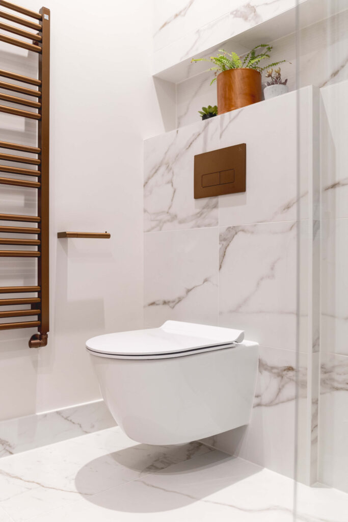 hugo-oliver-bathroom-sage-green-vessel-basin-copper-waste-white-worktop.jpg Wall-hung toilet with brushed copper flush plate, recessed display niche styled with plants, copper ladder towel rail and marble-effect wall tiles