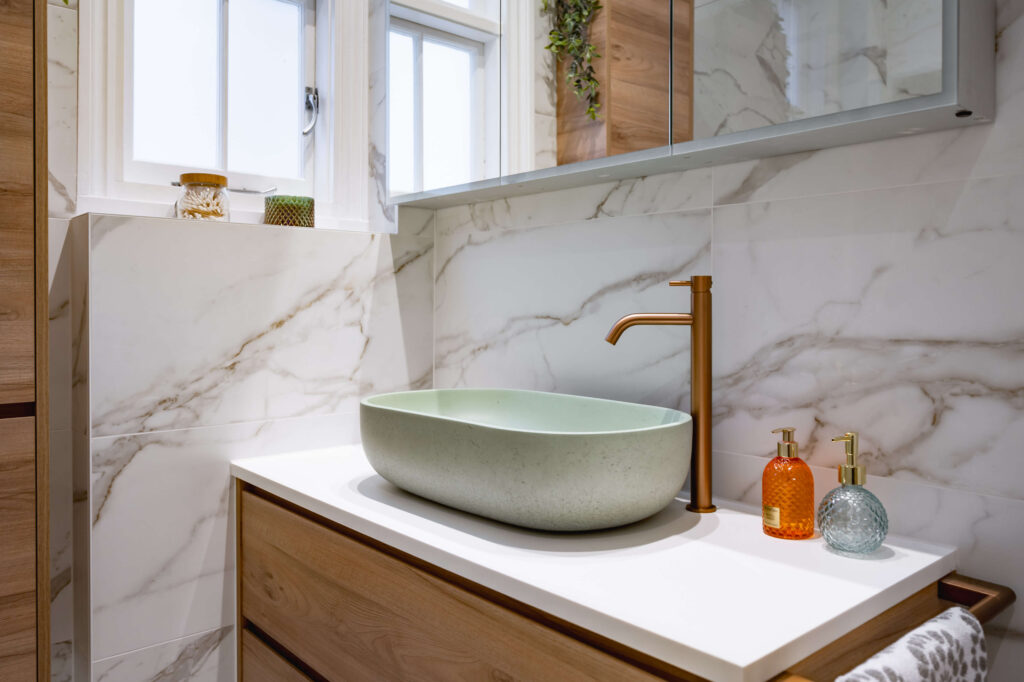 Close-up of sage green stone vessel basin with brushed copper tap, wood-effect tall storage column and marble-effect tiled walls