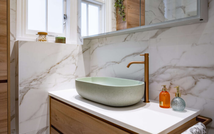 Close-up of sage green stone vessel basin with brushed copper tap, wood-effect tall storage column and marble-effect tiled walls